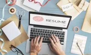 Dłonie piszące na laptopie z napisem "RESUME" i ikoną rakiety, otoczone materiałami do pakowania prezentów.