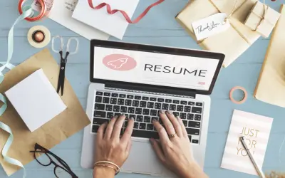 Dłonie piszące na laptopie z napisem "RESUME" i ikoną rakiety, otoczone materiałami do pakowania prezentów.