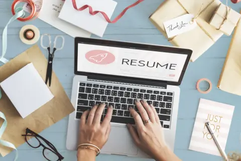 Dłonie piszące na laptopie z napisem "RESUME" i ikoną rakiety, otoczone materiałami do pakowania prezentów.