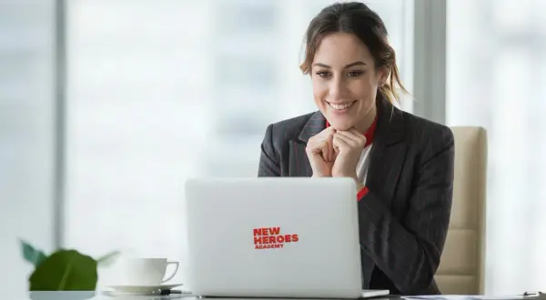 Uśmiechnięta kobieta w garniturze patrzy na laptopa z logo "New Heroes Academy".