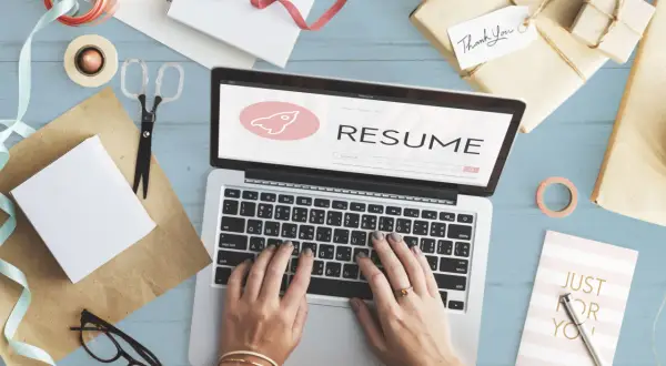 Dłonie piszące na laptopie z napisem "RESUME" i ikoną rakiety, otoczone materiałami do pakowania prezentów.